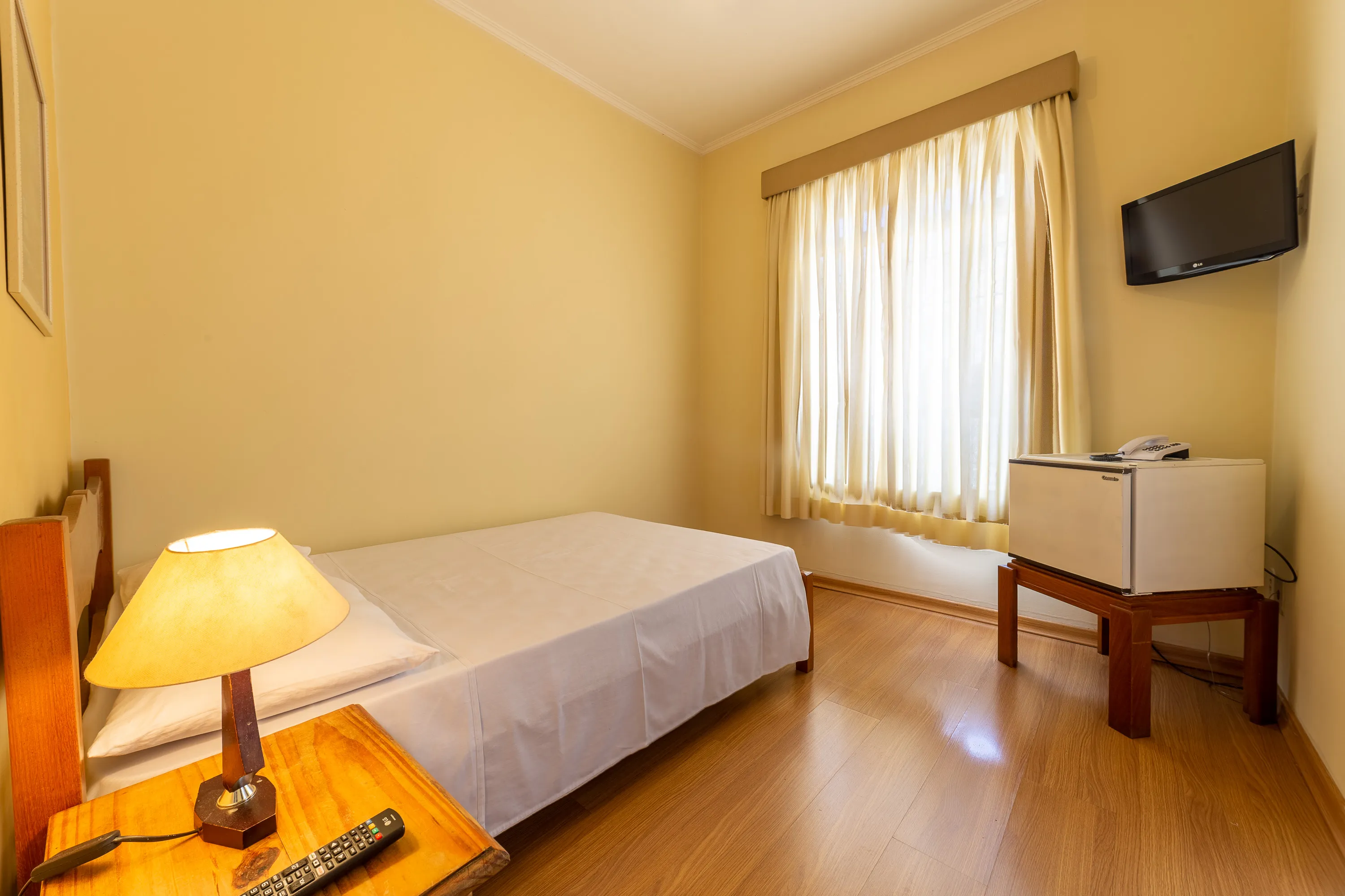 foto de quarto standard com ventilador, com uma cama de casal, em angulo mostrando mesa de cabeceira, tv e frigobar