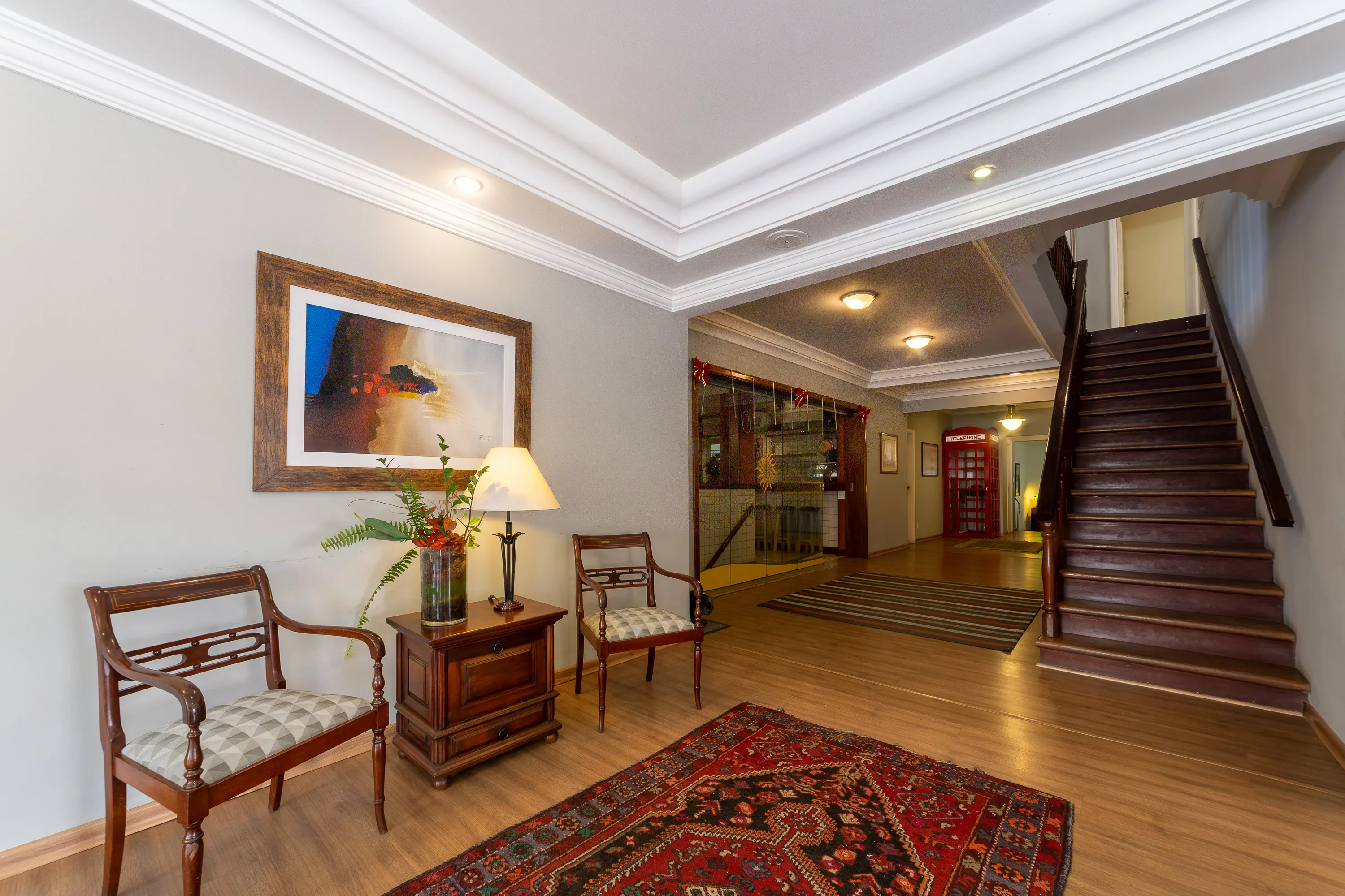 saguão principal do hotel com ambiente decorado com cadeiras e móveis em madeira maciça. é possível ver uma lanchonete com portas de vidro e uma escada que dá acesso aos quartos acima da recepção.
